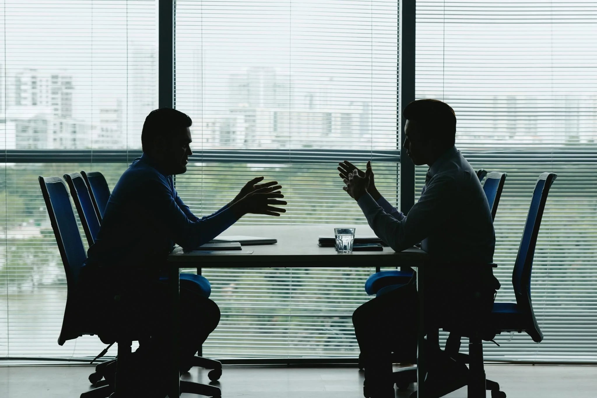 Two people sitting across from each other at a table in a modern office setting with large windows.